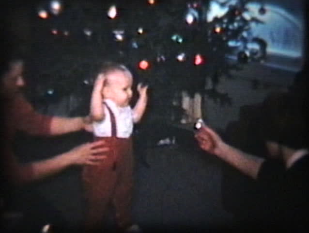 A cute baby boy with red suspenders tries to walk towards a shiny Christmas decoration.
