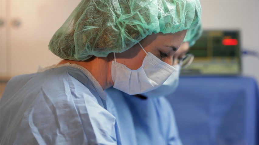 The camera dollies from a surgeon and surgical nurse busy in the operating room past two IV containers in the foreground.