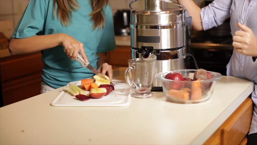 Two adolescent girls standing in a kitchen cut and add fruits and vegetables to a juicer to make a nutritious vitamin packed drink.