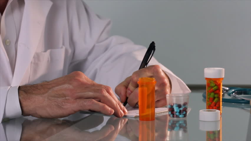 A pharmacist or doctor writes something on a prescription pad then pour some colorful caplets into a pill container.