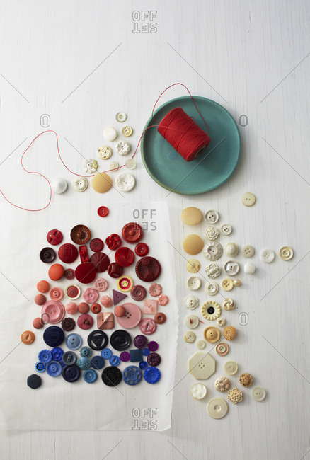 Still life of red, pink, blue and white buttons, aqua plate, red thread on wax paper and white wood surface