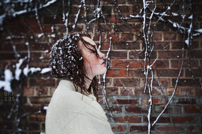 Young girl wrapped in beige pashmina seen in profile against brick wall in snowstorm.  Her hair is damp and covered in snow flakes