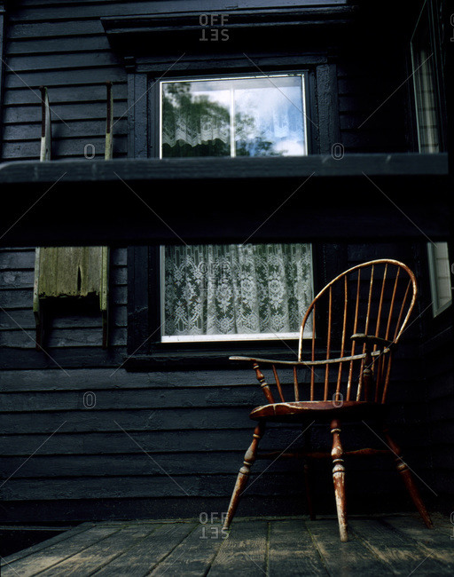 Old wooden windsor style chair on porch of dark gray house with lace curtain hanging in window photographed on RTE 1 in Maine