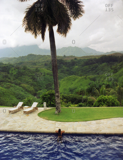 Girl leaning on side of pool with view of palm tree and mountains of Kauai in the distance