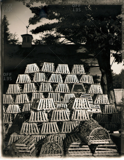 Black and white image of stacks of old wooden lobster pots with roof-line and weather vane in background