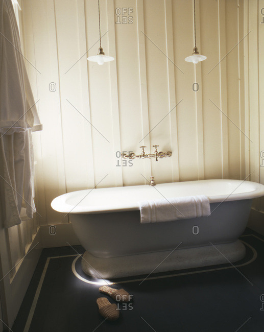 Old fashioned tub with slippers on gray floor and robe hanging on hook with two lamps over tub