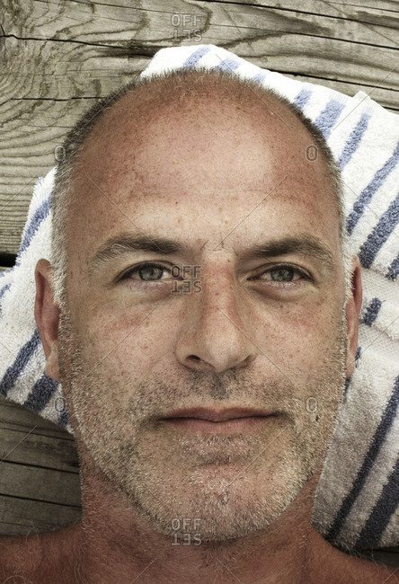 Close up portrait of bald, gray haired man resting his head on striped white and blue pillow on dock seen from above.