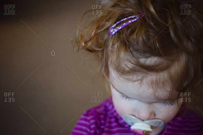 Close-up of curly red-haired female toddler with pacifier looking down.