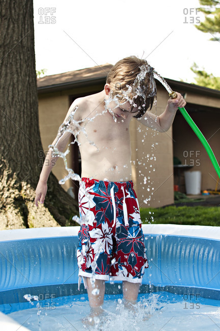 Young boy stands in swim pool while holding a running hose over his head