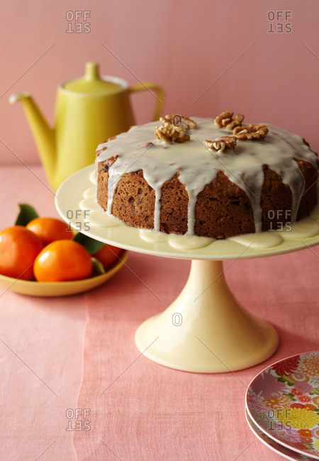 A whole glazed cake on a ceramic stand with floral pattern plates, mandarins and teapot.