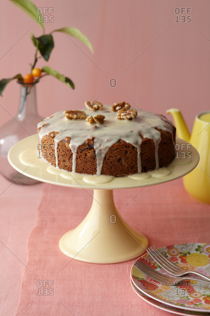 A whole glazed cake on a ceramic stand with floral pattern plates, fork and teapot.