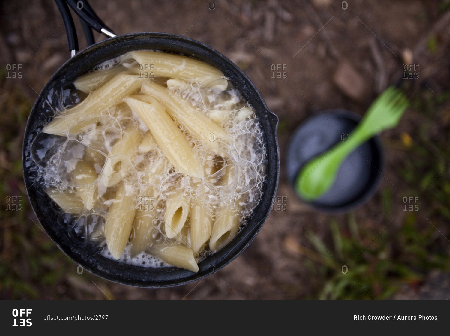 A camping stove boils over with pasta with a camping spoon in the background