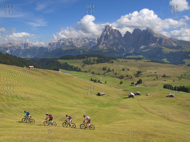 Four mountain bikers are riding downhill a grassy slope at Seiser Alm, with rock cliffs in the background