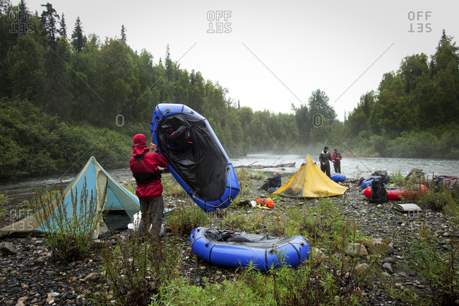 Four friends packing up camp by a river with tents and packrafts in the rain.