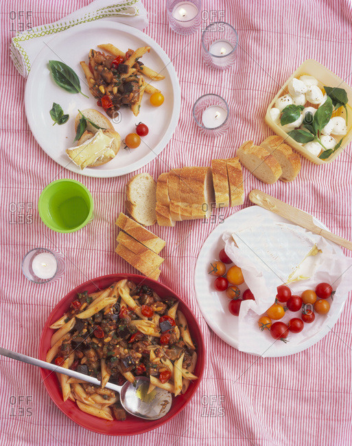 Picnic food spread of pasta salad, cherry tomatoes, a loaf of bread and mozzarella on a blanket with candles