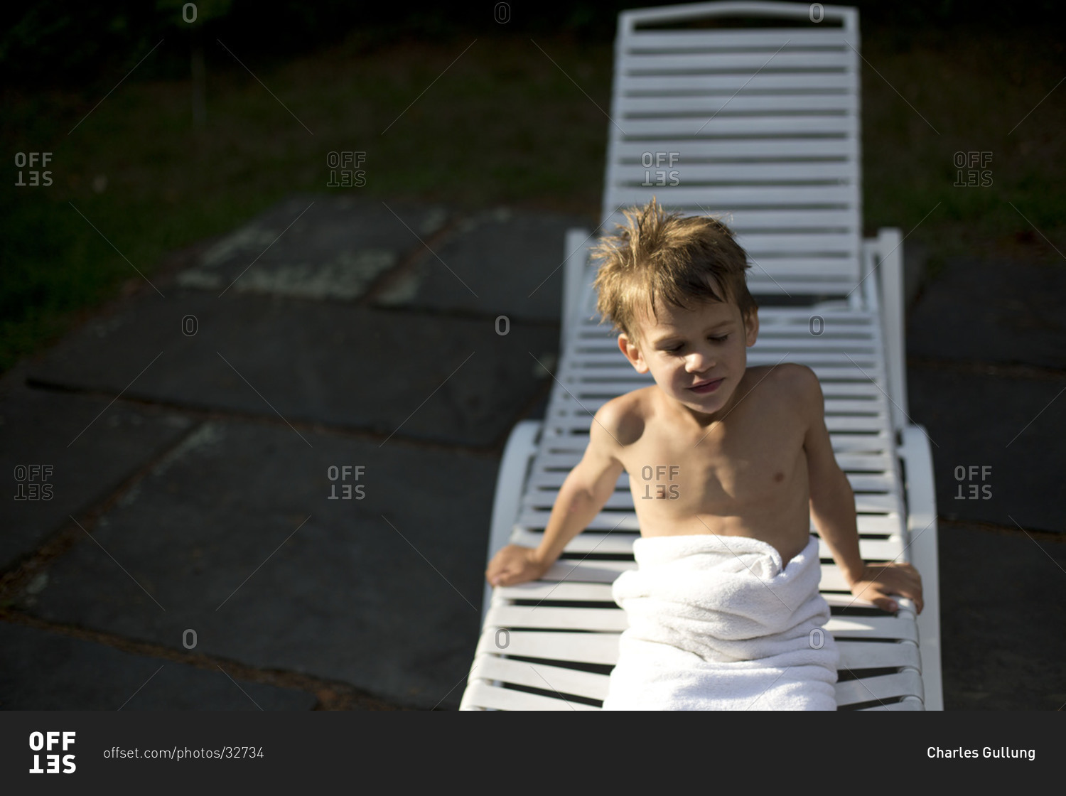 Little boy wrapped in white towel sitting on plastic deck chair.