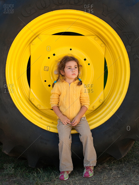 Young girl sitting on large yellow truck wheel and staring away pensively