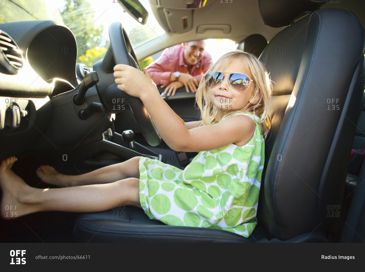 Portrait of little girl sitting in driver's seat of car, pretending to be old enough to drive as her smiling father watches on on a sunny summer evening in Portland, Oregon, USA