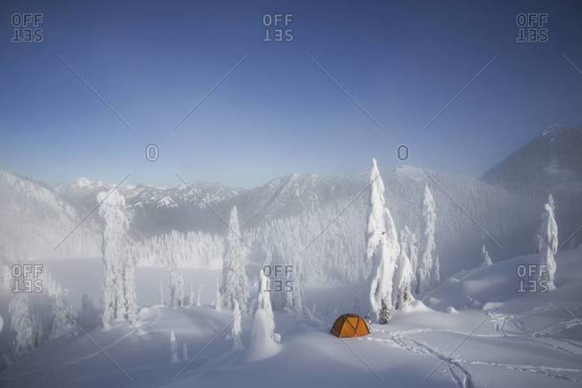 A bright orange tent among snow covered trees, on a snowy ridge overlooking a mountain in the distance.