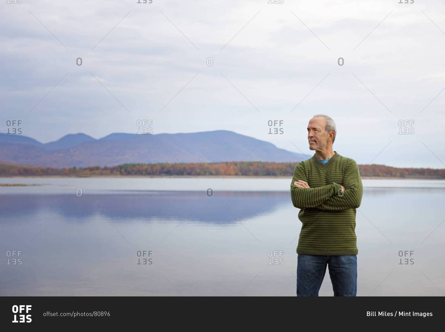 A man with arms folded by the water of a flat calm lake in the mountains