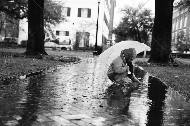 Young girl kneeling down while holding an umbrella picking a leaf from a puddle in the park