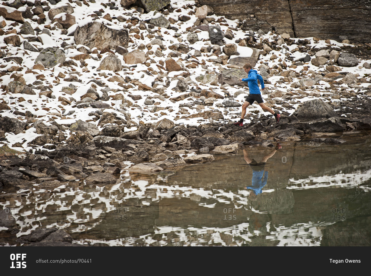 Athletic young man runs on a snowy mountain trail beside a lake with his reflection
