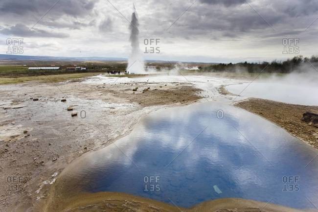 Blue water in geothermal pool with water spout from Strokkur Geysir visible in the distance, Geysir, near Reykjavik, Iceland, Polar Regions
