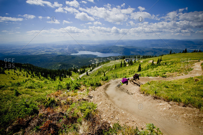 Two downhill mountain bikers chase each other down a winding trail on a sunny summer day.