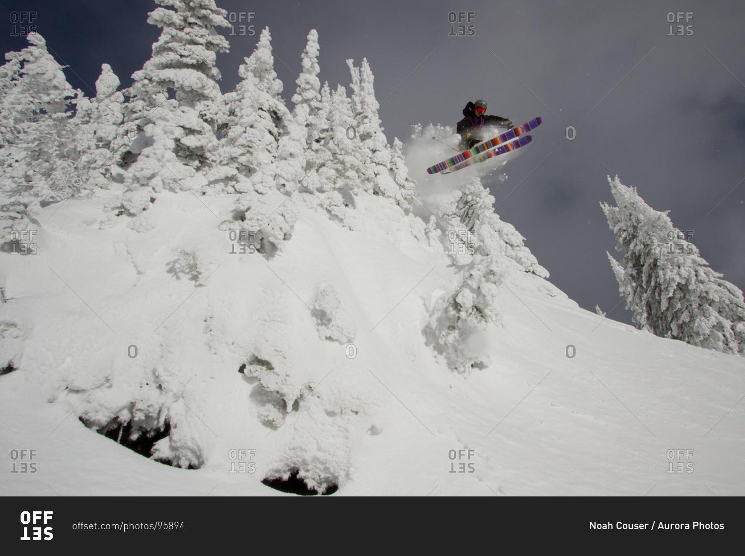 A skier catches air over a little cliff surrounded by snowcovered trees.