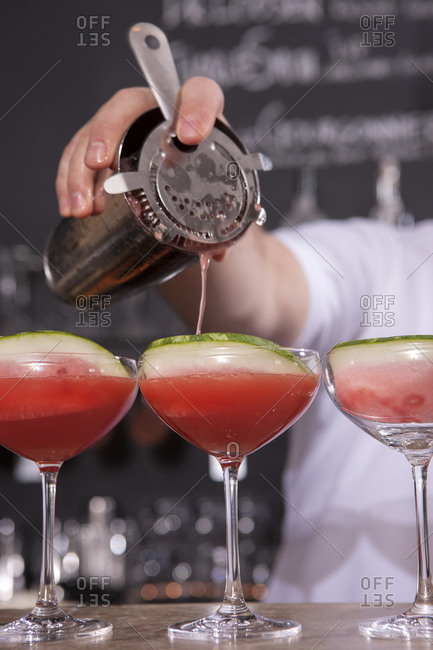 A bartender pours a drink into a martini glass containing a watermelon slice