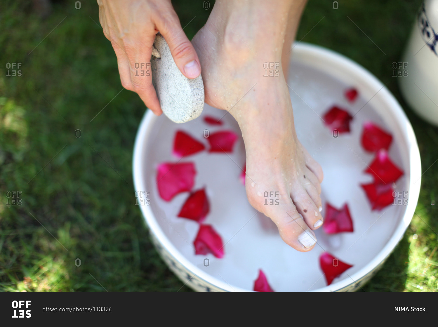 Woman taking an outdoor foot bath with rose leaves while exfoliating cracked skin on heel with pumice stone