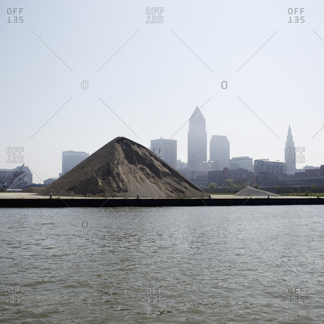 View over rippled water of a large mound of dirt and a city skyline