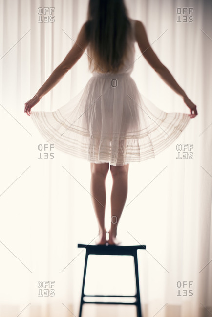 Young woman standing on a stool holding seam of her skirt in front of a white curtain