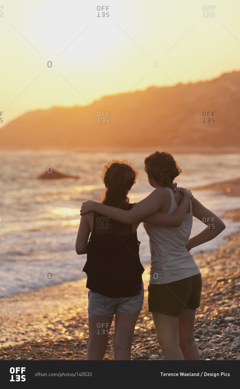 Two young women in an embrace watch the sunset while looking out over the ocean