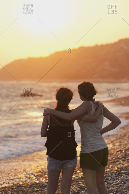 Two young women in an embrace watch the sunset while looking out over the ocean