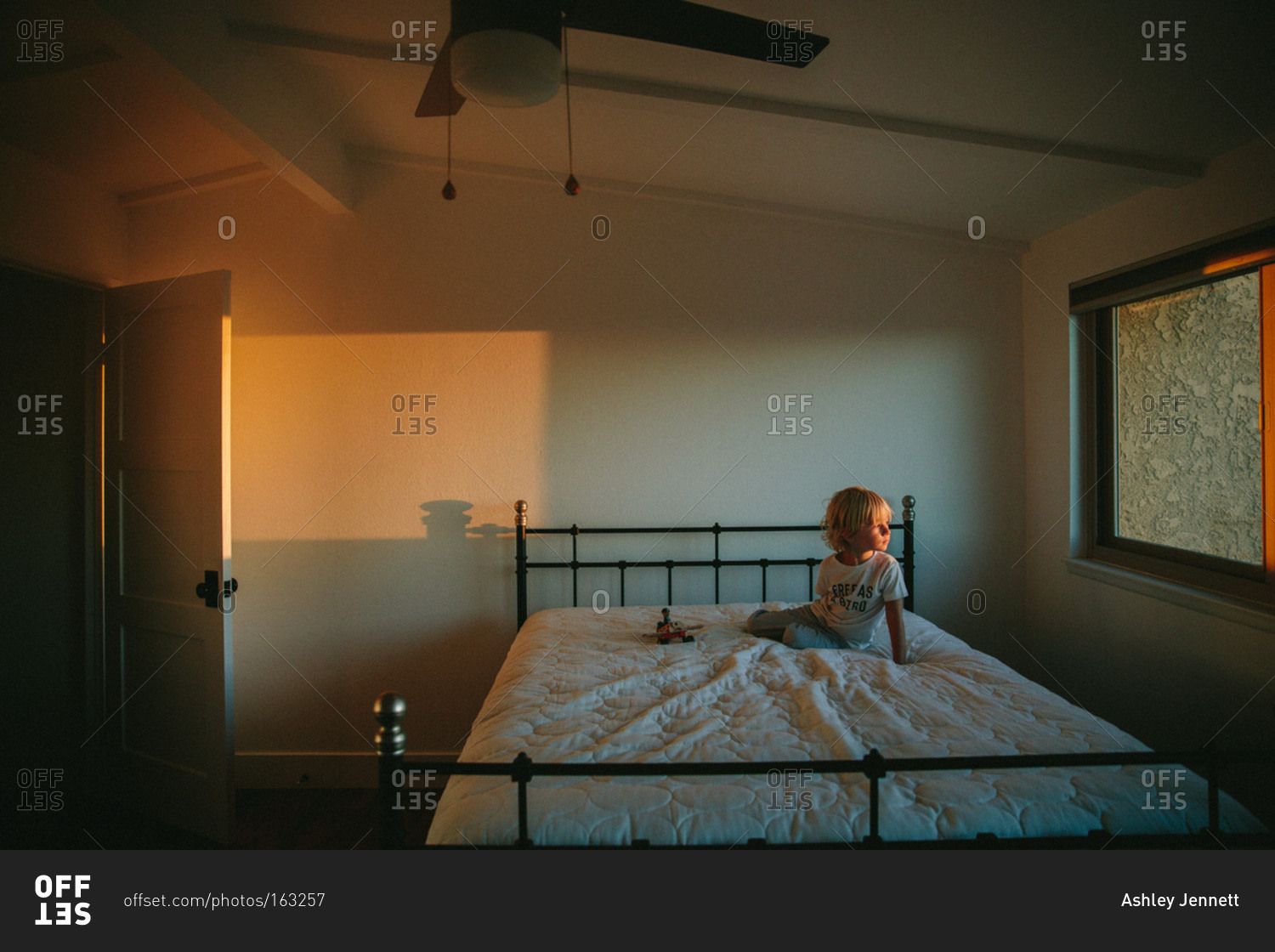 Young boy sitting on a bed and staring out of a window stock photo OFFSET