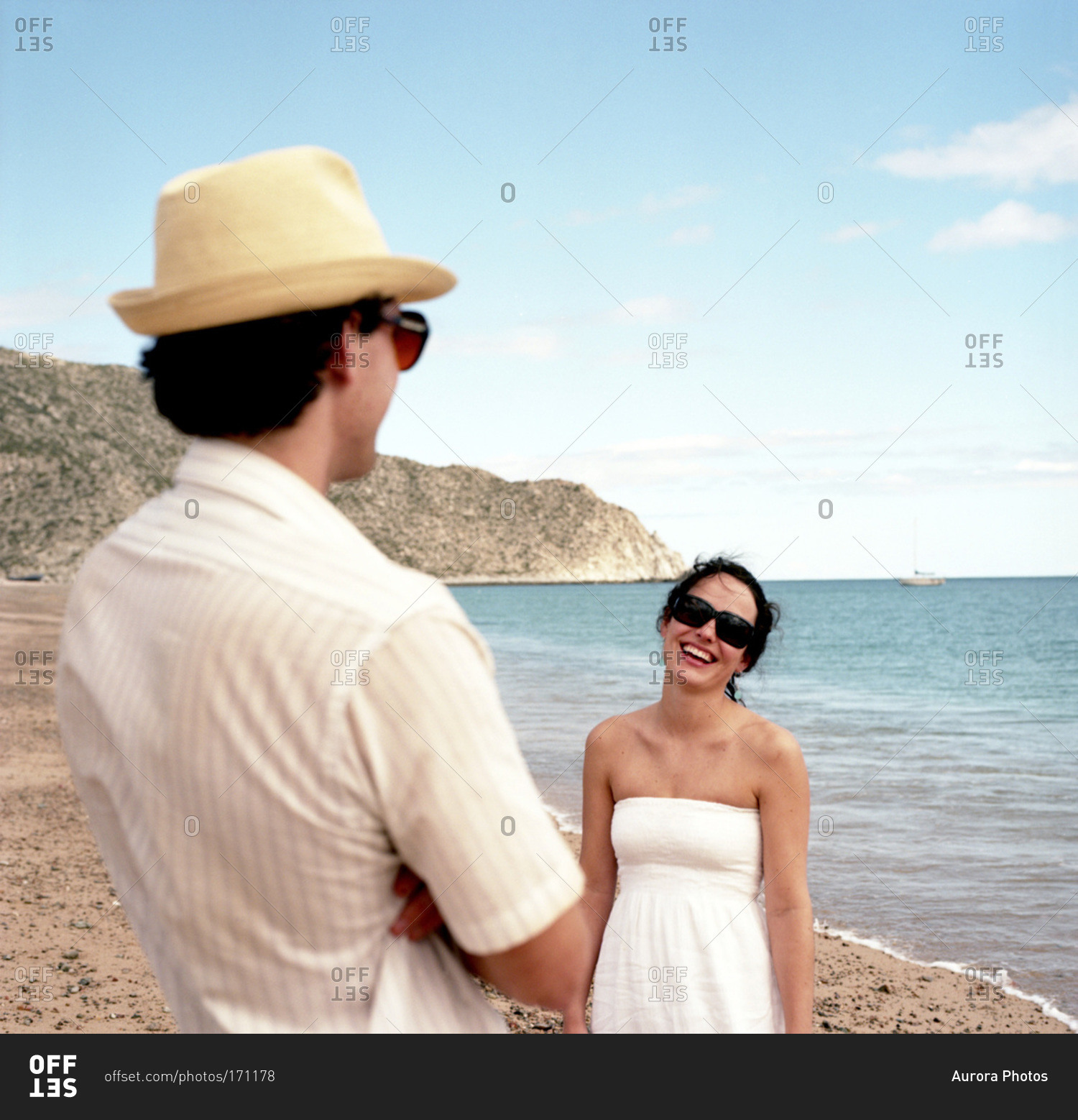 A woman smiles at her boyfriend as she walks down the beach on a sunny afternoon wearing a white sun dress