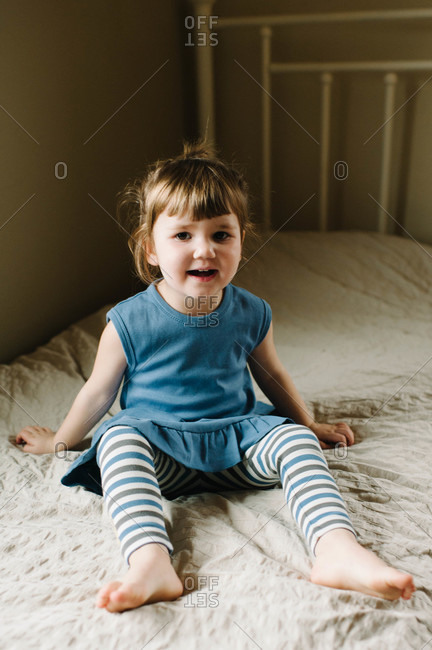 A little girl in a blue dress sits on her bed