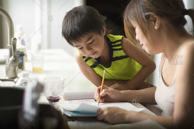A Japanese boy doing his Japanese homework in a kitchen as his mom looks on