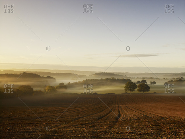 A ploughed field and view over surrounding undulating hills, at dawn with a mist rising from the land