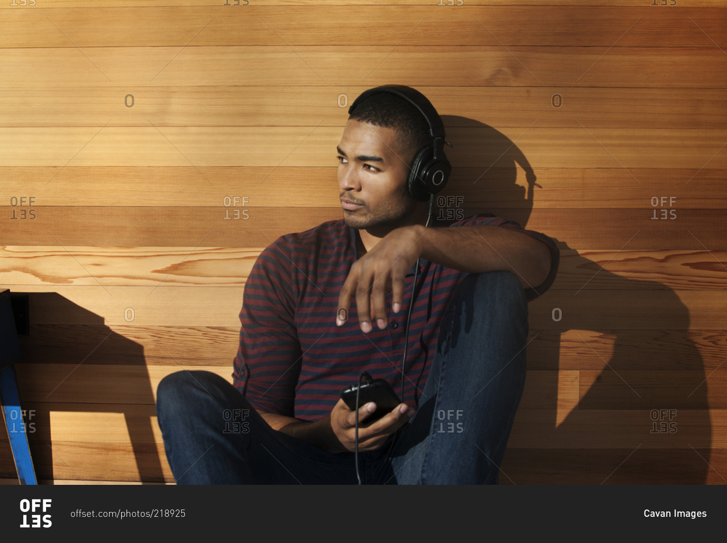 A young man leans against a wood paneled wall and wears headphones