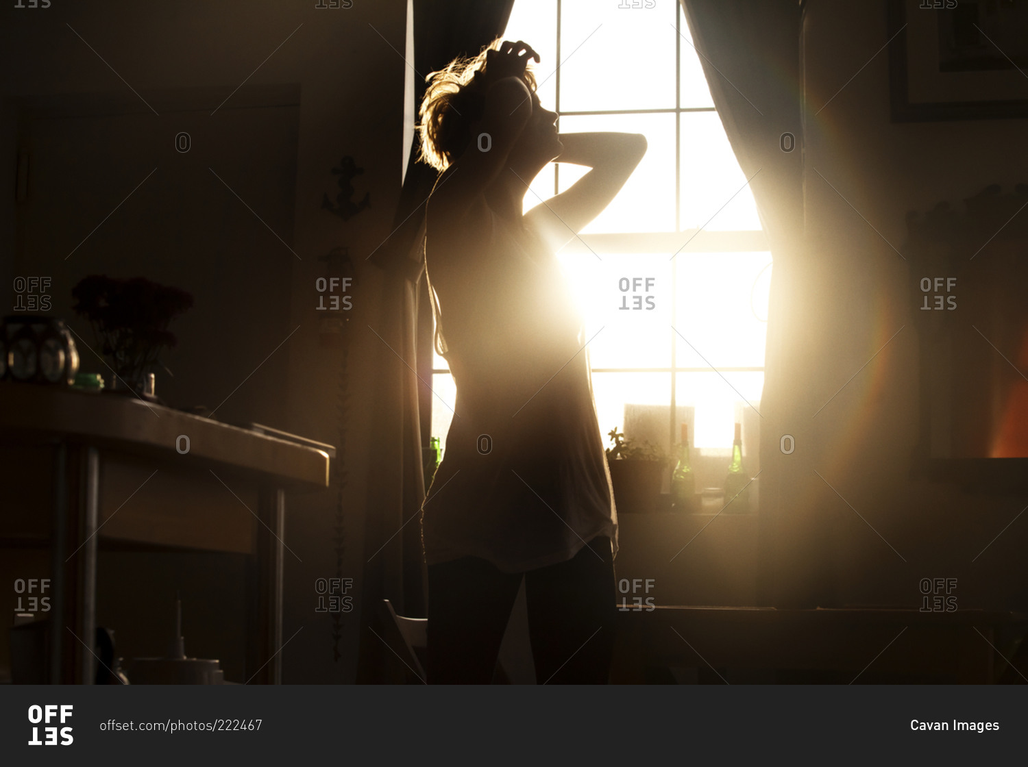 Backlit young woman standing by window with hands behind her head