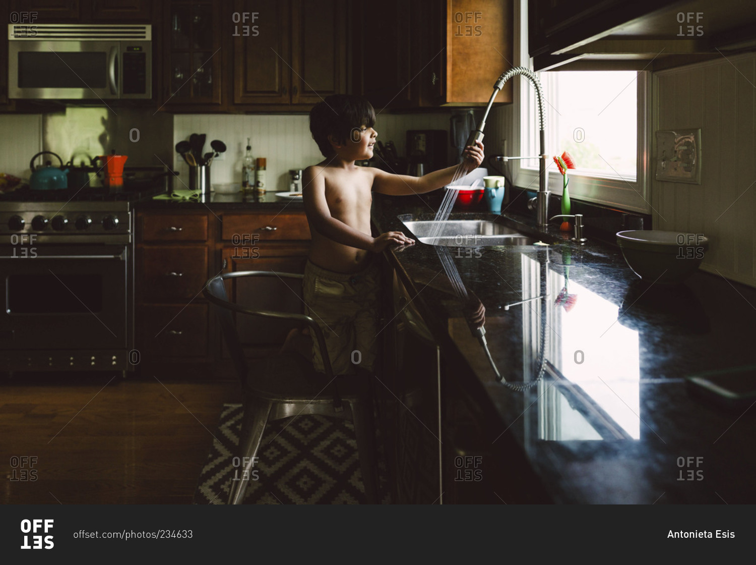 A boy kneels on a chair and rinses food in a sink