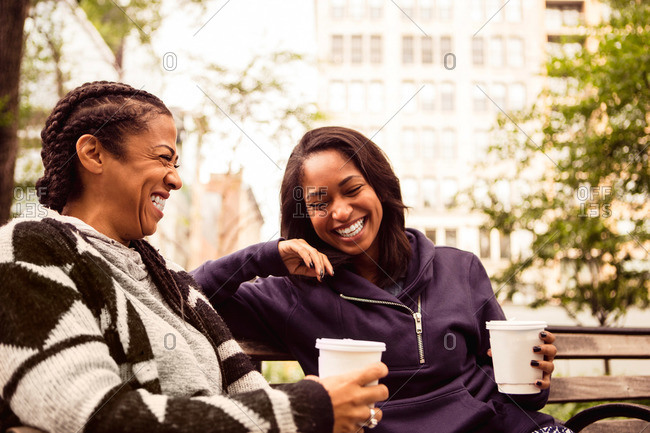 Two women laughing together while drinking coffee on a park bench