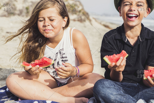 Siblings laugh while eating watermelon on the beach