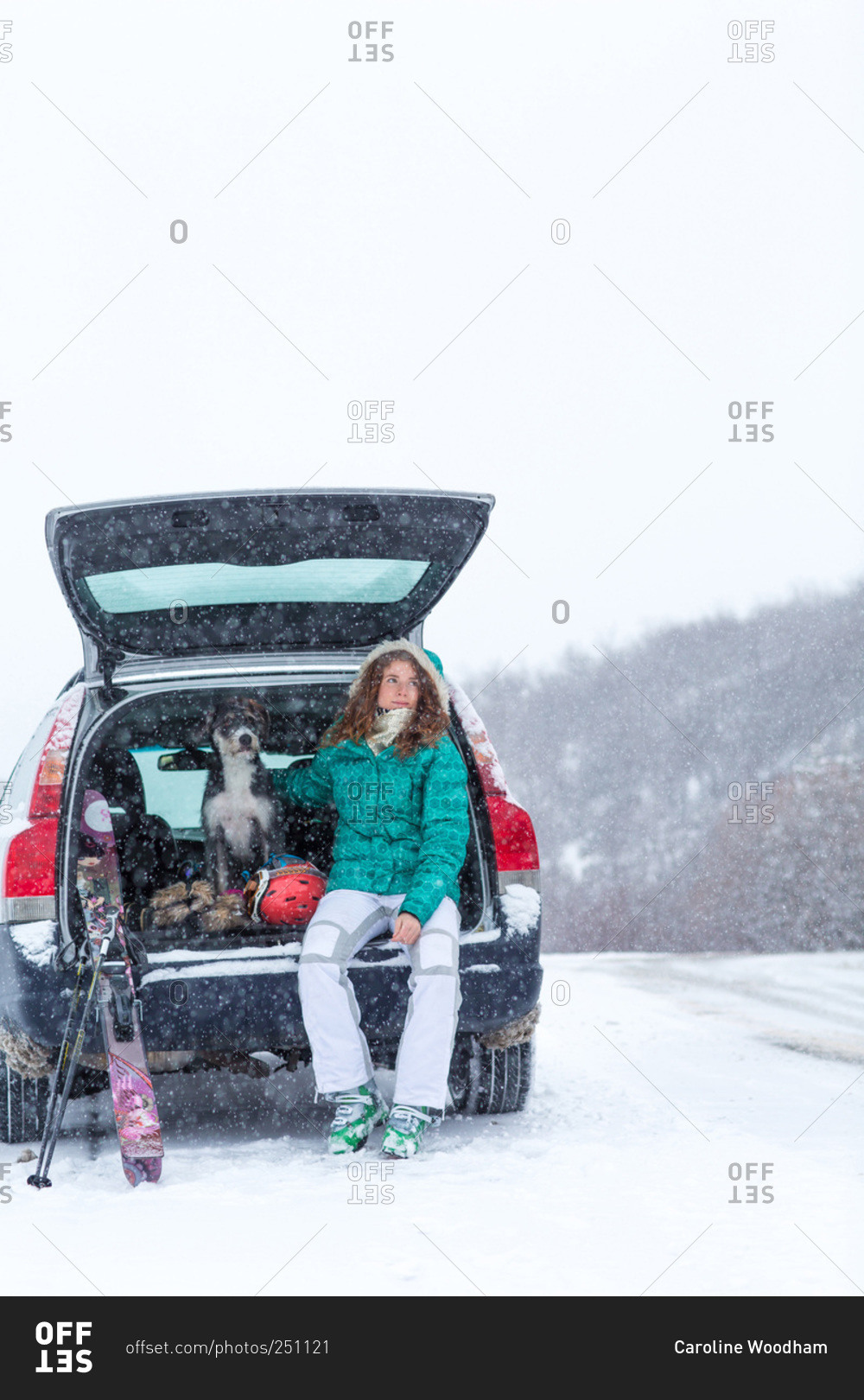 Young woman and her dog sit in their vehicle hatch with skis in snowstorm