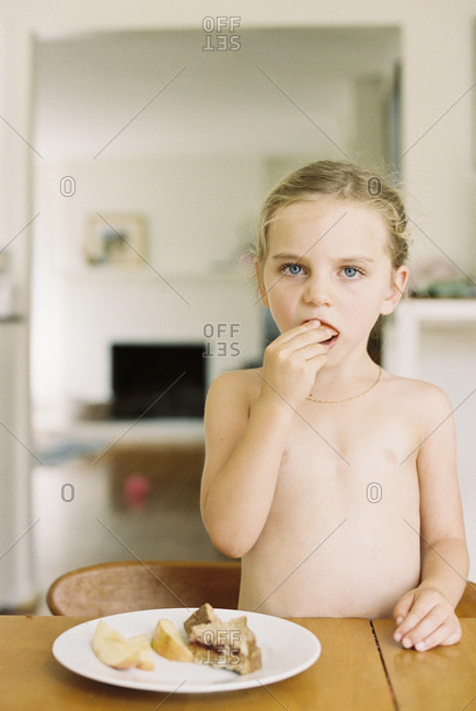 Nude young girl standing at a table, eating a sandwich and ...