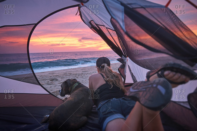 Woman and dog looking out tent at beach sunset