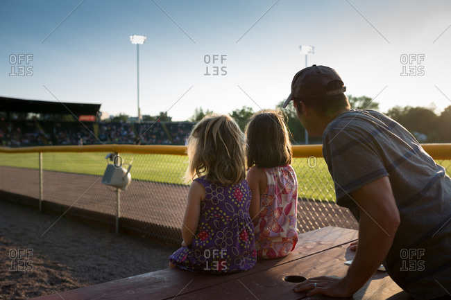 Back view of two young girls and their father watching a baseball game at picnic table