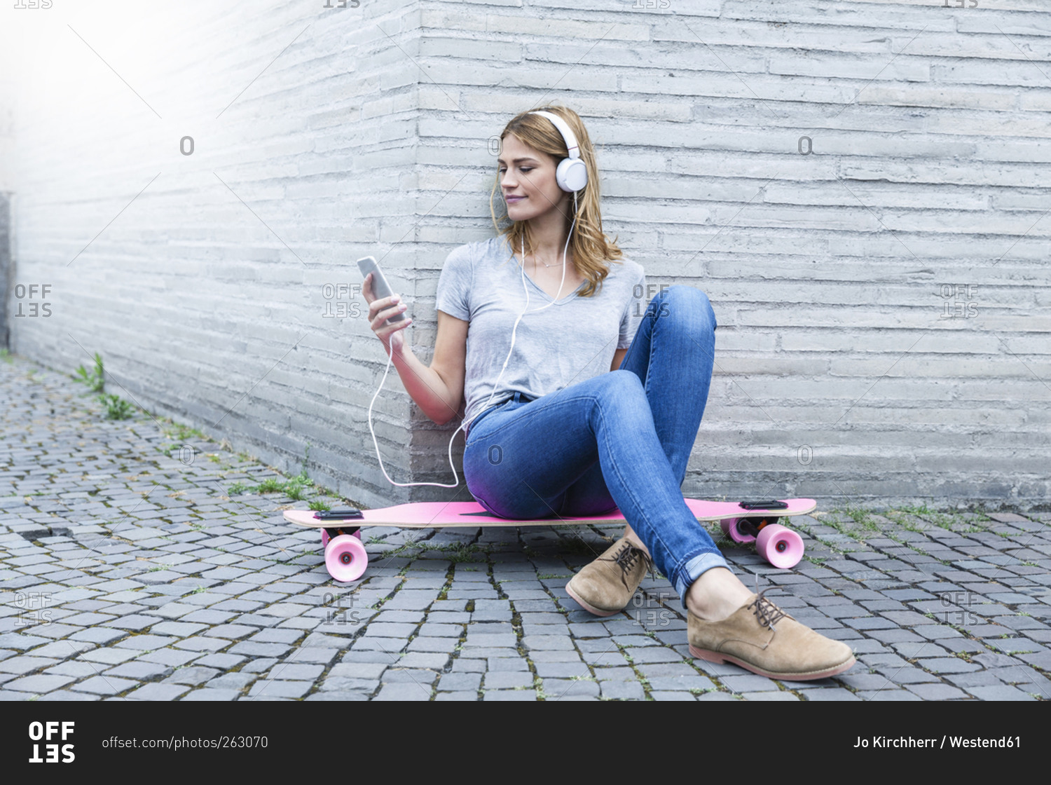 Young woman sitting on pink skateboard listening to music